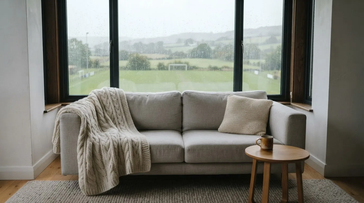 Ein Sofa mit Wolldecke steht vor einem grossen Fenster, draussen Regen und in der Ferne ein Fussballplatz.