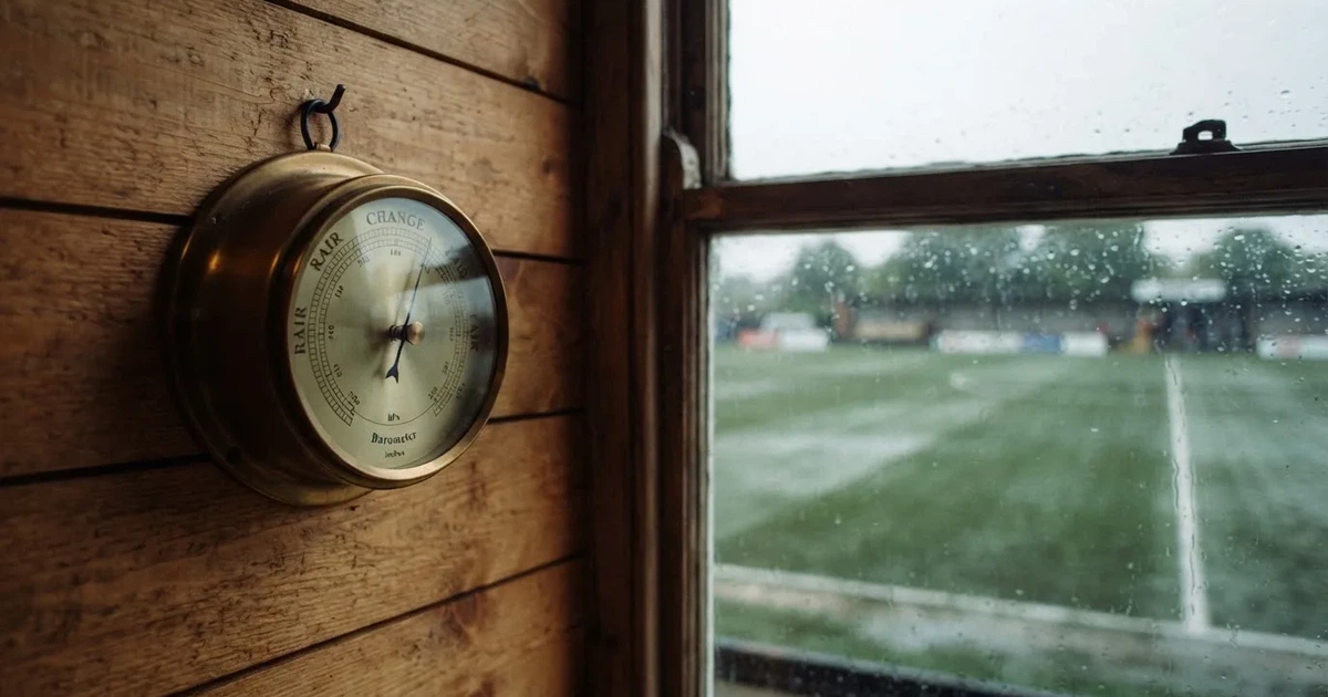Ein Wetterbarometer hängt an einer Holzwand neben einem Fenster, draussen liegt ein nasser Fussballplatz im Regen.