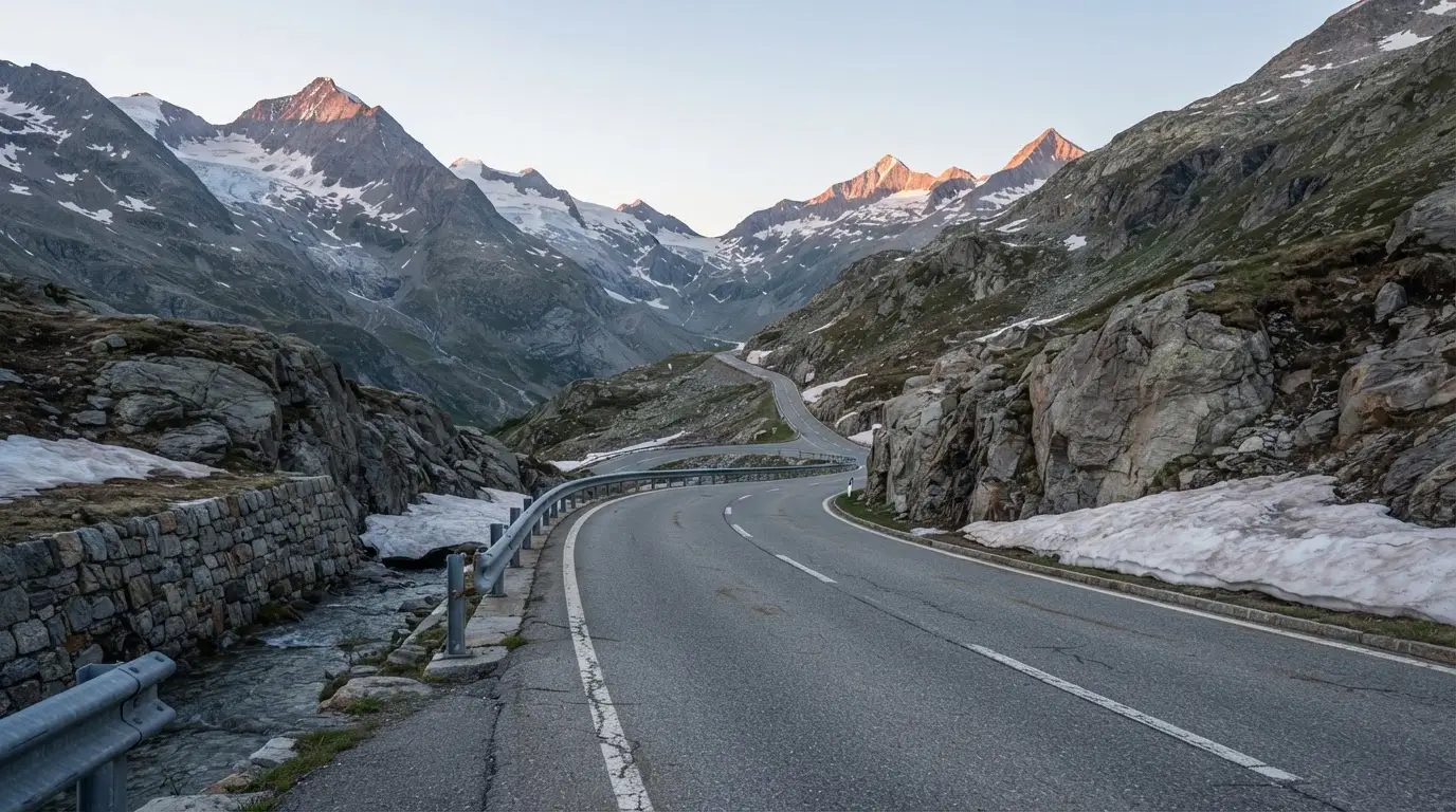 Ein Bergpass in den Schweizer Alpen mit leerer Strasse und Schneeresten am Rand im ersten Frühlicht.