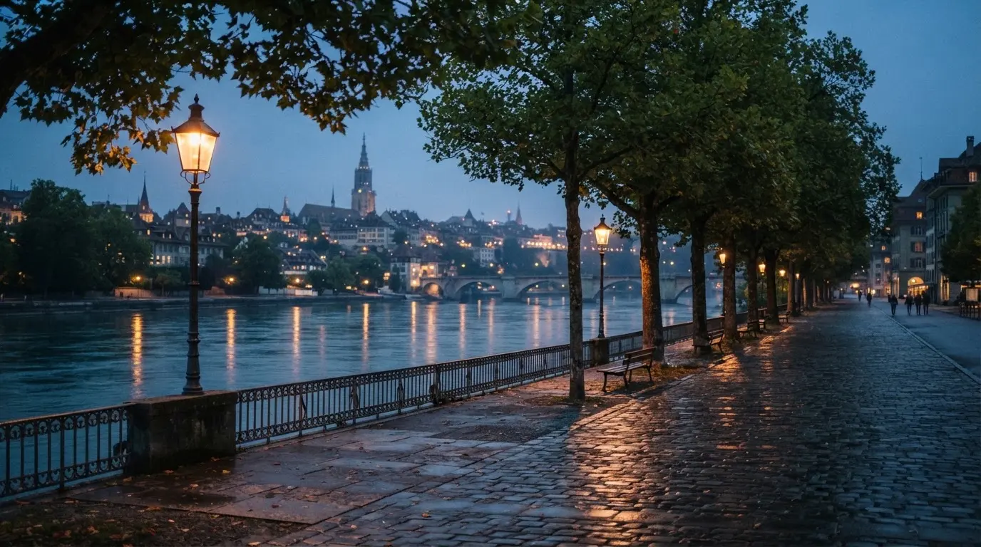 Eine Rheinpromenade mit hohen Bäumen am Abend, das Wasser spiegelt das Licht der ersten Laternen.