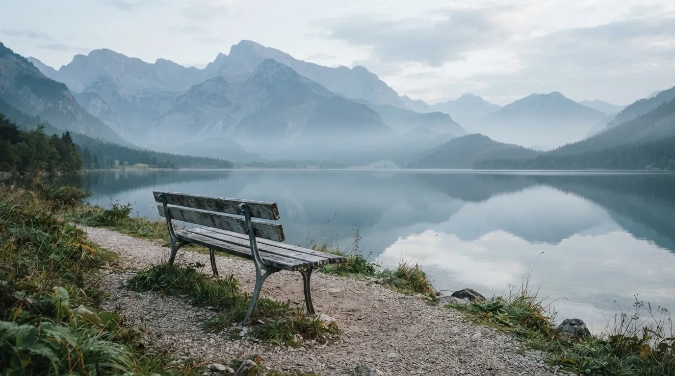 Eine Holzbank an einem Seeufer mit Blick auf eine Bergkette im Morgendunst, das Wasser ist spiegelglatt.